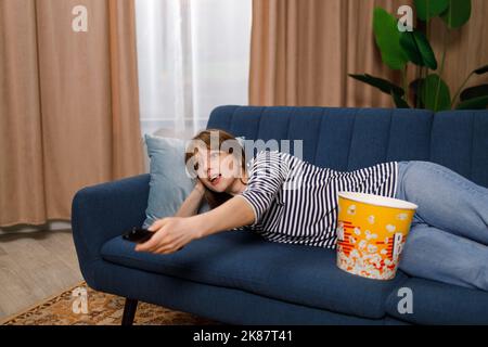 Frau liegt mit Popcorn auf dem Sofa, schaut fern und wechselt die Kanäle Stockfoto