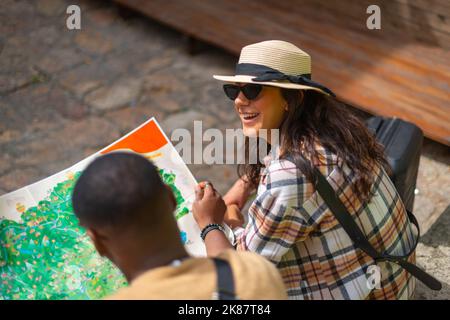 Ein cooles multiethnisches Paar sitzt und lacht, draußen, an einem sonnigen Tag Stockfoto