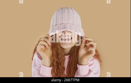 Lachende glückliche Mädchen trägt in rosa gestrickte Hut über die Augen auf beigem Hintergrund gezogen. Stockfoto