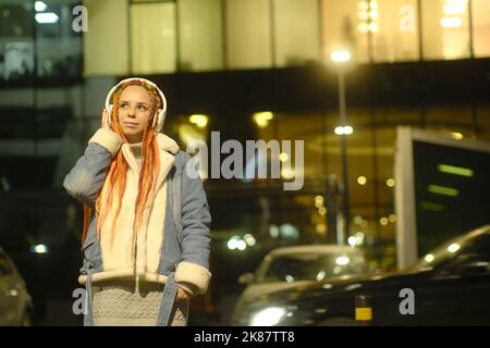 Content Frau, die nachts in der Stadt Musik hört. Positive junge Hündin mit langen orangefarbenen Dreadlocks und in Jeansjacke, die Musik im Headpho hört Stockfoto