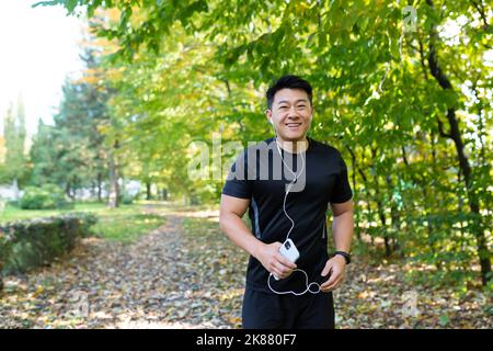 Porträt eines glücklichen Sportlers im Park, eines asiatischen Mannes, der läuft und Musik über Kopfhörer hört, die Online-Anwendung zum Hören von Radio und Podcasts nutzt, die Kamera anschaut und lächelt Stockfoto
