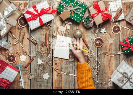 Weibliche Hand schreibt Frohe Weihnachten in einem Notizbuch auf einem Tisch mit Weihnachtsgeschenken und Dekoration Stockfoto