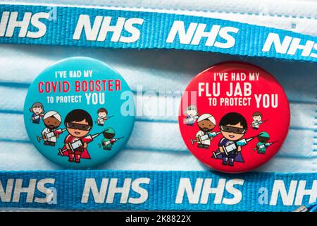 Farbenfrohe Covid 19 Booster- und Flu Jab-Abzeichen neben einem NHS-Trageband und einer chirurgischen Gesichtsmaske Stockfoto