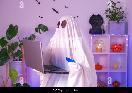 ghost of Halloween nutzt Laptop im Internet surfen, Online-Shops durchsuchen, Märkte. Ein Geist macht eine Bestellung vom Computer für Halloween. Ghost zahlt für den Online-Kauf per Kreditkarte. Neonlicht. Stockfoto