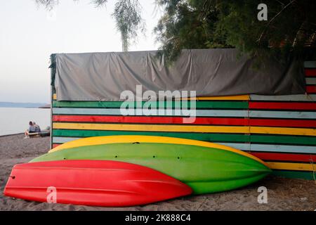 Kajaks / Kanus und bunte Schuppen an einem Sandstrand, Lesbos. September / Oktober 2022. Herbst. Zwei Personen sitzen. Stockfoto