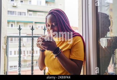 Junge afroamerikanische Frau, die Kaffee trinkt, sitzt am Küchenfenster Stockfoto