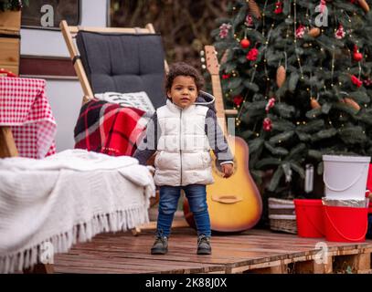 Kleines afroamerikanisches Mädchen in Winterweste, Jeans steht auf Holzpaletten in der Nähe des Trailers House. Der Hinterhof ist mit einem Weihnachtsbaum w dekoriert Stockfoto