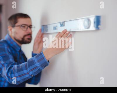 Junger Mann in blau Arbeitsanzug tun Reparatur Wohnung. Renovierungskonzept für das Haus. Kurzer Schuss eines hübschen bärtigen jungen Mannes, der zu Hause ein Level-Werkzeug hält Stockfoto