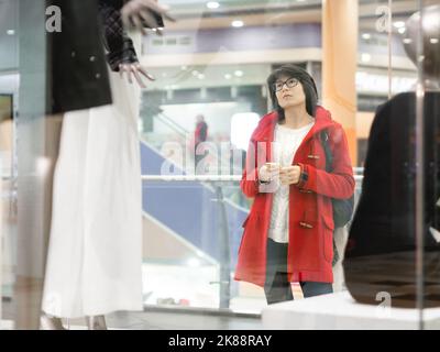 Frau starrt im Bekleidungsgeschäft auf Dummies. Lässiges Outfit an Schaufensterpuppen. Blick durch transparentes Schaufenster. Einkaufen im Einkaufszentrum. Einfache Kleidung Stockfoto