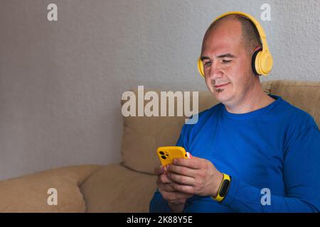 Ein lächelnder, kahler Kaukasier mit einem blauen Langarm-T-Shirt hört Musik von seinem Handy mit gelben Kopfhörern, die auf einem Sofa sitzen. T Stockfoto