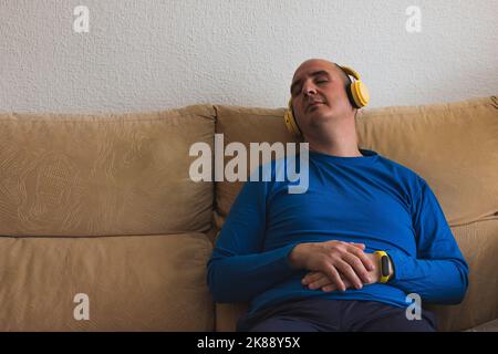 Ein kahler Kaukasusmann, der in einem blauen Langarm-T-Shirt gekleidet ist, schläft auf einem Sofa, während er gelbe Kopfhörer trägt. Stockfoto