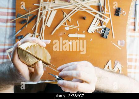 Hände des Mannes, der überschüssigen Klebstoff von Sperrholzdetails für Schiffsmodell mit Gebrauchsmesser entfernt. Prozess des Bauens Spielzeug Schiff, Hobby, Handwerk. Tabelle mit Stockfoto