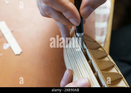 Hände des Mannes, der überschüssigen Klebstoff von Sperrholzdetails für Schiffsmodell mit Gebrauchsmesser entfernt. Prozess des Bauens Spielzeug Schiff, Hobby, Handwerk. Tabelle mit Stockfoto