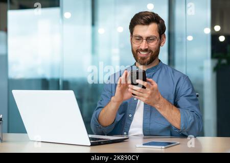 Erfolgreicher und glücklicher Geschäftsmann mit Bart arbeitet im modernen hellen Büro, männlicher Chef verwendet Laptop, Geschäftsinhaber im lässigen Hemd hält Telefon, durchsucht Internetseiten Stockfoto