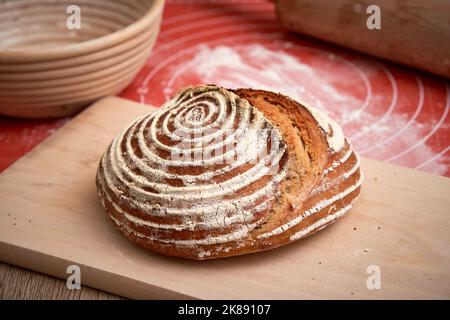 Hausgemachtes Sauerteig-Brot, das zu Hause gebacken wird. Schöne Kruste des Brotes. Cane Banneton im Hintergrund Stockfoto