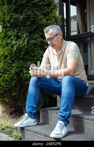 Trauriger, frustrierter älterer Mann, der in einer Brille schielt, benutzt Smartphone und sitzt auf einer Treppe in der Nähe des Eingangs. Technophobe brauchen Hilfe Stockfoto