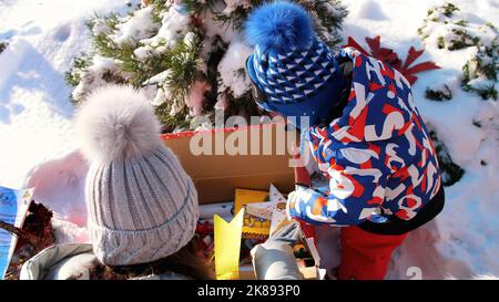 Winter, frostig, verschneit, sonniger Tag. Glückliche Familie mit kleinen Kindern Auspacken Sie Kartons mit Geschenken unter dem Weihnachtsbaum, im Freien. Hochwertige Fotos Stockfoto