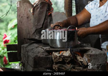 Nahaufnahme der Hände einer lateinischen Frau, einer braunhäutigen Bauernin, die einen Eisentopf auf einem traditionellen Ofen hält, der häufig zum Kochen auf einem Kessel verwendet wird Stockfoto