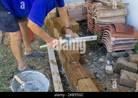 Bilder von den Händen eines alten maurers, während er mit Zement eine niedrige Mauer aus Tuffziegeln baut. Mann bei der Arbeit Stockfoto