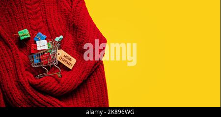 Weihnachtsbanner zum Einkaufen. Warenkorb mit vielen Geschenkboxen auf rotem Strick und gelbem Hintergrund Stockfoto