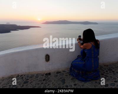 Die Dame nimmt den Sonnenuntergang über Thirasia auf ihrem Handy von einem Aussichtspunkt in Fira auf. Die griechische ägäische Kykladen-Insel Santorin. Stockfoto