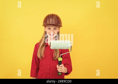 Fröhliches Teenager-Kind im Baumeister-Harthut mit Farbrolle an der gelben Wand Stockfoto