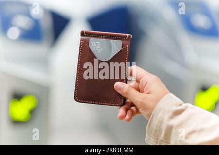 Weibliche Hand hält einen Kartenhalter aus Leder mit silberner Dubai Metro Nol Card an einem Bahnhof, automatische Tore im Hintergrund. Kontaktloser elektronischer Tarif. Stockfoto