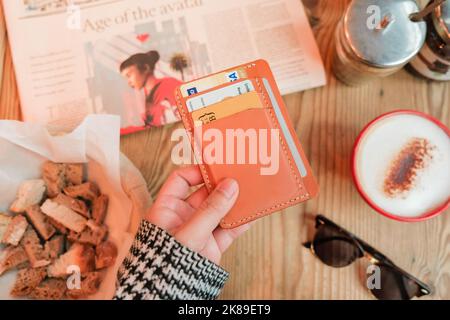 Die weibliche Hand hält einen Kartenhalter aus braunem Leder mit Ausweisen und Kreditkarten über einem Holztisch mit Kaffee, Brotkorb, Zeitung und Sonnenbrille. Stockfoto