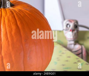 Ein Stillleben eines leuchtend orangefarbenen Kürbisses (Cucurbita pepo) auf einem Tisch mit einem Skelett, das auf einem Stuhl dahinter sitzt. Stockfoto