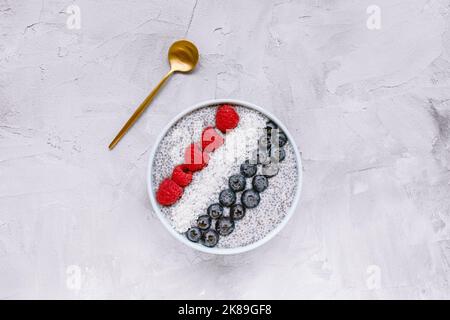 Über Nacht Chiasamen-Pudding mit frischen Heidelbeeren, Himbeeren und Kokosflocken in einer Schüssel mit goldenem Löffel Copy Space, Draufsicht. Frühstück, Superfood und veganes Food-Konzept. Grauer Hintergrund Stockfoto