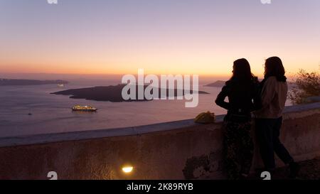 Zwei Frauen genießen den Sonnenuntergang über der Insel Thirasia von einem Küstenpfad in Fira aus. Die griechische ägäische Kykladen-Insel Santorin. Stockfoto