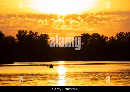 Fischer in einem Boot bei Sonnenuntergang auf dem Fluss auf dem Hintergrund des Waldes in der Ukraine in der Stadt Dnepr, goldener Sonnenuntergang auf dem Dnepr Fluss Stockfoto