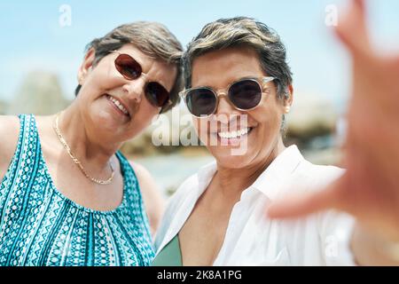 Age hat uns den Spaß nicht genommen. Zwei reife Freunde stehen zusammen und posieren für ein Selfie während eines Tages am Strand. Stockfoto