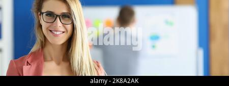 Lächelnde Geschäftsfrau posiert im Büro mit in vorzeigbarem rosa Kostüm, Kollege auf dem Hintergrund Stockfoto