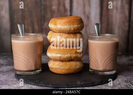 Zwei Gläser Milch mit Kakao mit vier glasierten Donuts auf einem Steinteller. Stockfoto