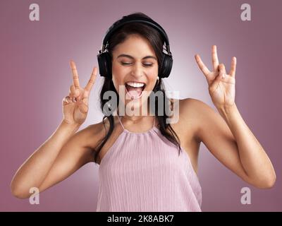 Sei ein Cupcake in einer Welt von Muffins. Eine schöne junge Frau zeigt das Friedenszeichen, während sie Musik hört, die Kopfhörer gegen ein Rosa trägt Stockfoto