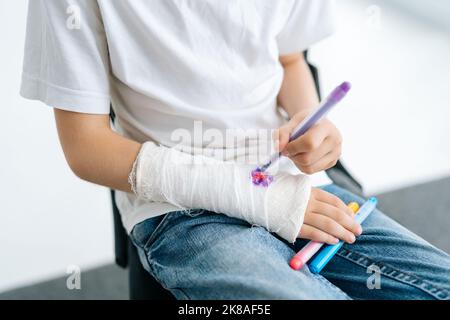 Nahaufnahme eines nicht erkennbaren kleinen Mädchens, das ein niedliches Bild auf einen gebrochenen Arm zeichnet, der in einen weißen Gipsverband eingewickelt ist, auf einem weißen isolierten Bild Stockfoto