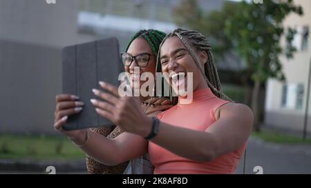 Zwei junge schwarze Frauen, die Selfie mit Tablet machen. Afroamerikanische Freundinnen mit geflochtenen Frisur halten Gerät im Freien Stockfoto