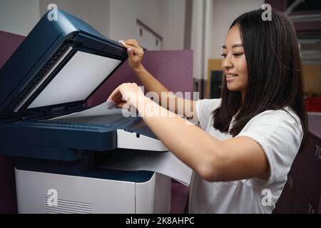 Lächelnde asiatische Frau arbeitet im modernen Büro mit Drucker Stockfoto