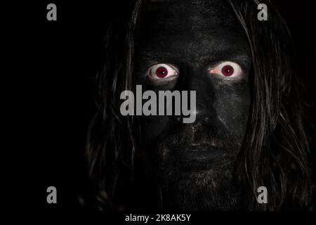 Ein beängstigendes Gesicht eines Mannes im Dunkeln. Lange Haare, schwarzes Gesicht und rote Augen. Schwaches Licht mit dunklem Hintergrund. Horrorkonzept. Stockfoto