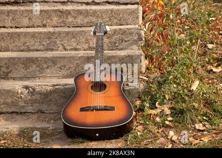 Alte braune Gitarre liegt im Park auf den Stufen im Herbst in der Sonne, Gitarre Stockfoto