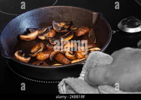 Braten von Kastanienpilzen mit einem Mann, der eine gusseiserne Pfanne mit einem Geschirrtuch in der Hand hält, auf einem Elektroherd Stockfoto