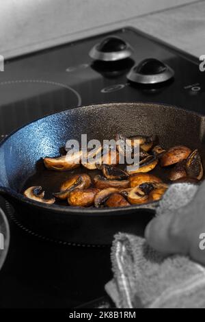 Braten von Kastanienpilzen mit einem Mann, der eine gusseiserne Pfanne mit einem Geschirrtuch in der Hand hält, auf einem Elektroherd Stockfoto