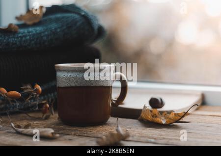 Große Tasse Cappuccino auf vintage braunem Hintergrund. Herbst, Herbstblätter, heiße, dampfende Tasse Kaffee und ein warmer Schal auf Holztischhintergrund. Meere Stockfoto