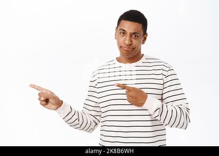 Skeptischer schwarzer Mann, der verärgert aussehe, mit den Fingern nach links auf das Logo oder Banner zeigend, im gestreiften Hemd auf weißem Hintergrund stehend Stockfoto