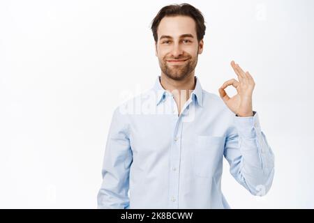Selbstbewusst lächelnder Geschäftsmann, Verkäufer zeigt okay, OK Zeichen in Genehmigung, wie smth, empfiehlt Unternehmen, steht über weißem Hintergrund Stockfoto
