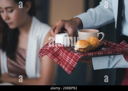 Der Kellner hält Teller mit Brot und einer Tasse Kaffee, während er die Gäste in einem Restaurant bedient. Stockfoto