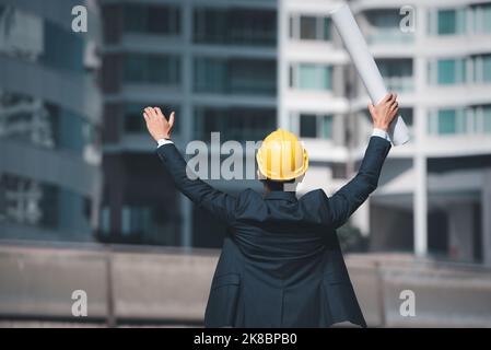 Der Architekturingenieur hob die Hände und hielt den Bauplan, um den Erfolg der Baustelle zu zeigen. Stockfoto