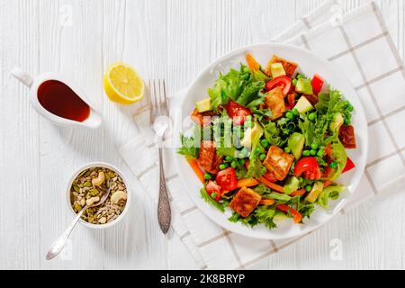 Tofu-Salat mit Avocado, knackigen Salatblättern, grünen Erbsen, Tomaten, gelber Paprika in weißer Schüssel mit Sesamöl-Dressing auf weißem Tisch, flach Stockfoto