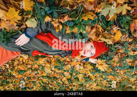 Draufsicht auf eine verträumte Frau mit herbstgelben Blättern in der Hand in orangefarbenem Strickhut und auf Blättern im Park liegendem Scarfl Stockfoto